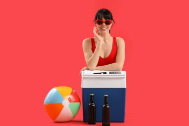 Portatif buzdolabı ve kırmızı arka planda soğuk bira şişeleri olan genç bir kadın.