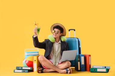 Dizüstü bilgisayarı ve kokteyli olan genç işadamı sarı arka planda yaz tatilini bekliyor.
