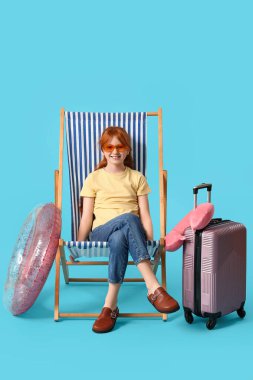Güverte sandalyesinde bavulu ve mavi arka planda şişirilebilir yüzüğüyle oturan tatlı kız. Seyahat kavramı