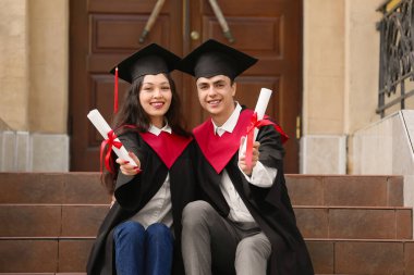 Üniversitenin yanındaki merdivenlerde oturan diplomalı birkaç mutlu mezun öğrenci.