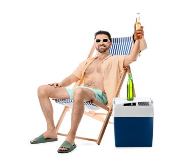 Portatif buzdolabı ve soğuk bira şişesiyle beyaz arka planda izole bir şekilde güverte sandalyesinde oturan mutlu genç adam.