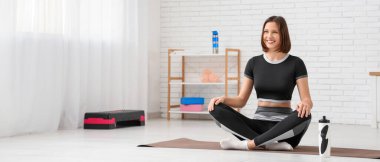 Spor salonundaki spor paspasında oturan genç bir kadın.