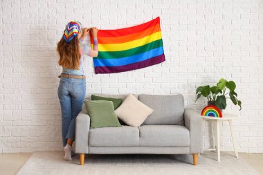 Oturma odasındaki beyaz tuğla duvara LGBT bayrağı asan genç bir kadın.