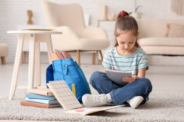 Sırt çantalı, defterli ve kalemli şirin bir kız yerde oturup evde ders çalışıyor.