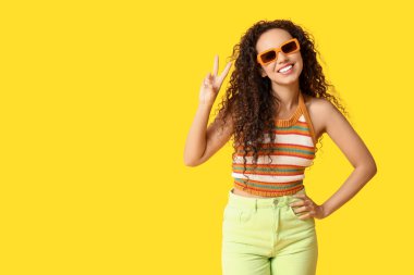 Güneş gözlüklü mutlu Afro-Amerikalı kadın sarı arka planda zafer jesti gösteriyor.