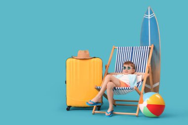 Güverte sandalyesinde oturan şirin küçük çocuk şişme top, bavul ve mavi arka planda sörf tahtasıyla. Seyahat kavramı