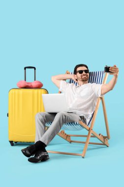 Genç iş adamı dizüstü bilgisayarı güverte sandalyesi ve bavuluyla mavi arka planda selfie çekiyor. Yaz tatili kavramı