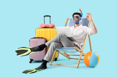 Dizüstü bilgisayarı olan genç işadamı güverte sandalyesinde ve mavi arka planda plaj aksesuarlarında iyi görünüyor. Yaz tatili kavramı