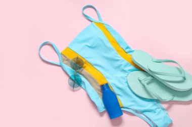 Şık bir mayo, parmak arası terlik, güneş gözlüğü ve pembe arka planda bir şişe güneş kremi.