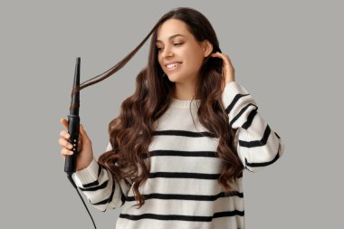 Beautiful young woman with curling iron on grey background