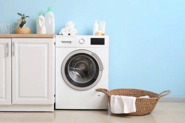 Mavi duvarın yanında sepeti ve dolabı olan modern çamaşır makinesi. Ev çamaşır odasının içi.