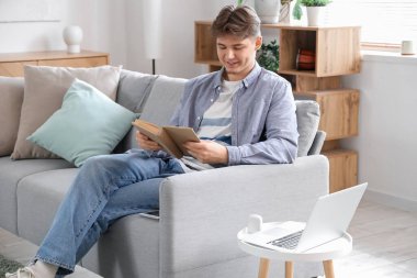 Kitabı olan bir erkek öğrenci evdeki kanepede ders çalışıyor.