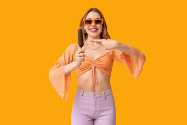 Mutlu gülümseyen genç kadın sarı arka planda çikolata kaplı dondurmayı işaret ediyor.