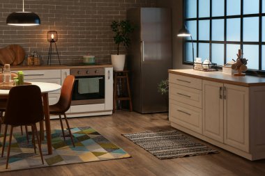 Parlayan lambalar, beyaz tezgahlar, fırın ve akşam yemeği masası olan mutfağın içi.