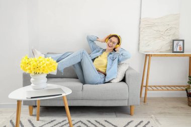 Kulaklıklı genç bir kadın evdeki kanepede müzik dinliyor.