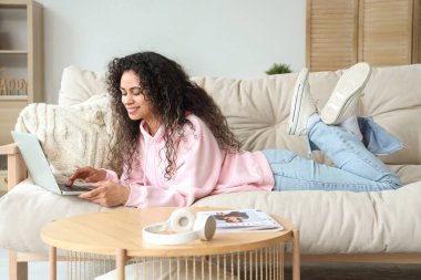 Genç Afro-Amerikalı kadın evdeki kanepede dizüstü bilgisayar kullanıyor.