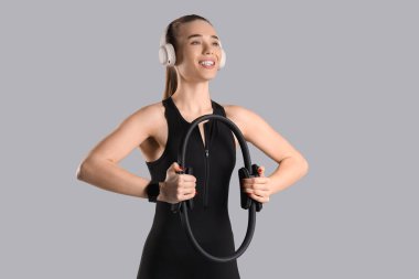 Gri arka planda pilates ringi olan kulaklıklı genç bir kadın.