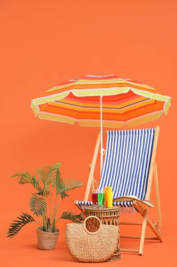 Çizgili iskemle, şemsiye, ev bitkisi ve turuncu arka planda plaj aksesuarları.
