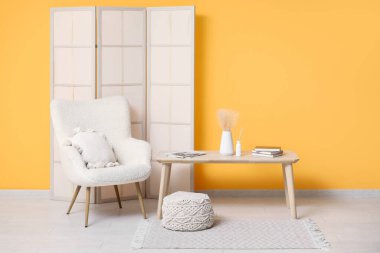 Sarı duvarın yanında katlanan ekran, koltuk, puf ve masa olan odanın içi.