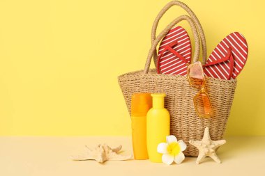Hasır çanta, güneş kremi şişeleri, deniz yıldızları ve sarı arka planda plaj aksesuarları, yakın plan.