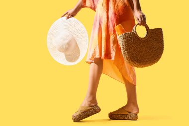 Sarı arka planda plaj şapkası ve çantası olan genç bir kadın.