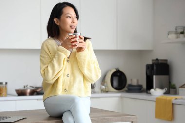 Bir fincan lezzetli çayla mutfakta oturan güzel Asyalı kadın.