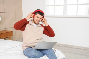 Kulaklık ve dizüstü bilgisayarlı genç sakallı adam yatak odasında müzik dinliyor.