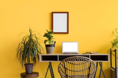 Ofiste sarı duvarın yanında dizüstü bilgisayarı ve bitkileri olan modern bir iş yeri.