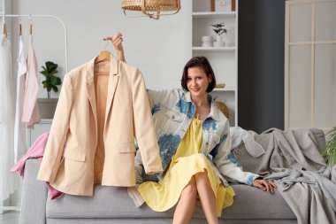 Genç, hayal kırıklığına uğramış bir kadın ceketini tutuyor ve evdeki farklı kıyafetlerin yanında kanepeye oturuyor.