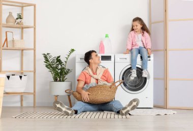 Genç baba ve kızı çamaşır sepetiyle banyoda çamaşır makinelerinin yanında oturuyorlar.