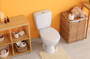 Tuvaletin içinde turuncu duvarın yanında klozet, raf ve sepet var.