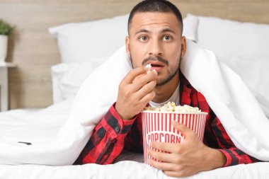Patlamış mısırlı genç adam yatakta film izliyor.