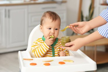 Anne, bebeğini evdeki yüksek sandalyede yeşil püre ile besliyor.