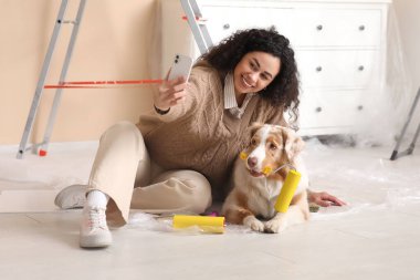 Yeni evinde tamir edilirken selfie çeken, bigudili ve şirin Avustralyalı çoban köpeği olan mutlu Afro-Amerikan ressam kadın.