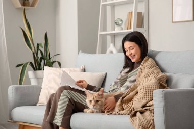 Battaniyeli güzel genç bir kadın ve evdeki kanepede kitap okuyan sevimli bir Maine Rakun kedisi.