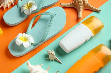 Parmak arası terlikli kompozisyon, plumeria çiçekleri, deniz kabukları ve renk arka planında güneş kremi şişeleri, yakın plan