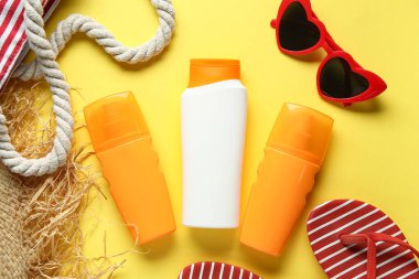 Çantalı kompozisyon, şapka, güneş kremi şişeleri ve sarı arka planda güneş gözlüğü.