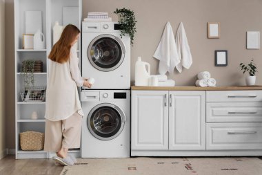 Çamaşır deterjanı banyodaki çamaşır makinesine koyan genç bir kadın.