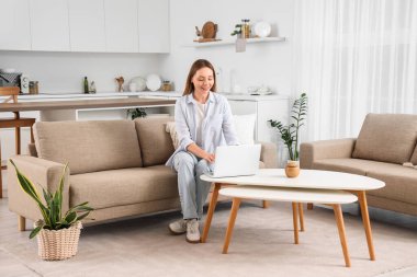 Kanepede oturan ve ışık odasında dizüstü bilgisayar kullanan güzel genç bir kadın.