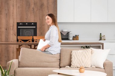 Oturma odasında dizüstü bilgisayarıyla kanepede oturan güzel genç bir kadın.