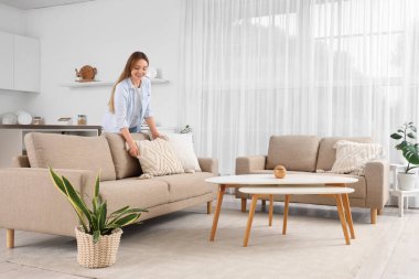 Güzel genç bir kadın, oturma odasındaki koltukta yastığı ayarlıyor.