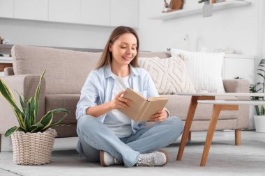 Genç ve güzel bir kadın yerde oturuyor ve oturma odasında kitap okuyor.