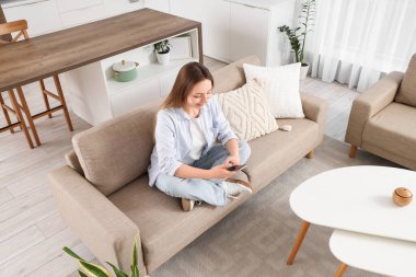 Güzel genç bir kadın kanepede oturuyor ve oturma odasında cep telefonu kullanıyor.