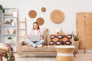 Güzel genç bir kadın kanepede oturuyor ve oturma odasında dizüstü bilgisayar kullanıyor.