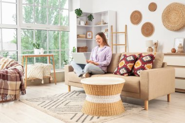 Güzel genç bir kadın kanepede oturuyor ve oturma odasında dizüstü bilgisayar kullanıyor.