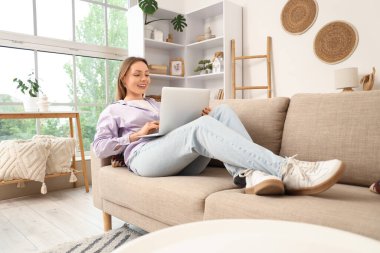 Güzel genç bayan kanepede uzanıyor ve oturma odasında dizüstü bilgisayar kullanıyor.