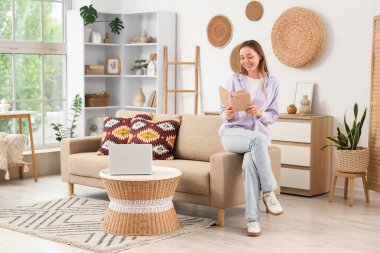 Genç ve güzel bir kadın kanepede oturuyor ve oturma odasında kitap okuyor.