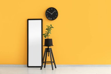 Sarı duvarın yanındaki taburede büyük bir ayna ve ev bitkisi var.