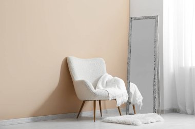 Oda içindeki bej duvarın yanında yer aynası ve koltuk var.