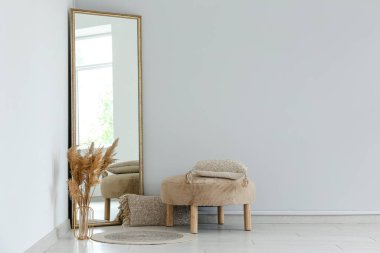 Zemin aynası, vazo içinde otlar ve beyaz duvarın yanında puf var.